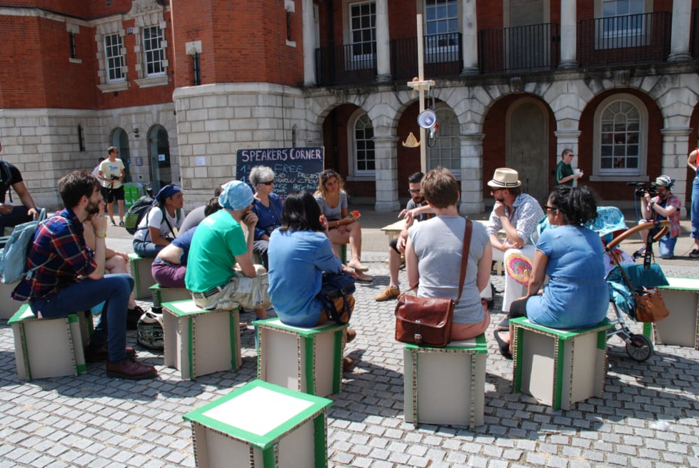 People sitting on cardboard stools in an outdoor space, all facing inwards as one person at the front is speaking.