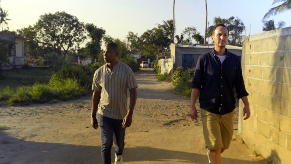 Two men walking together down a sandy road, with low buildings behind them