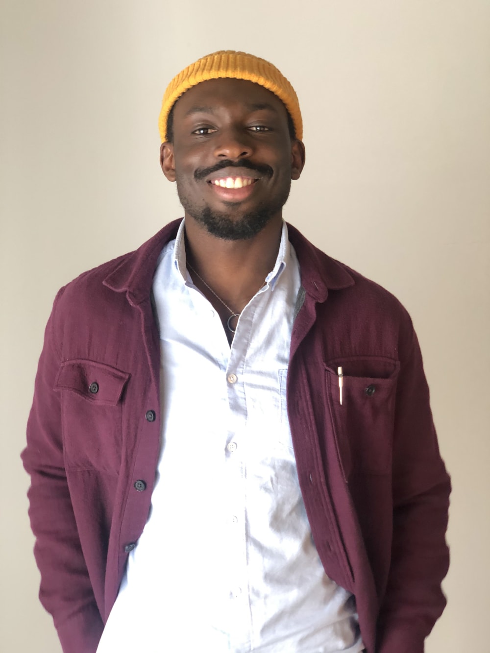 Photo of Olu smiling into the camera. He's wearing a yellow beanie, white shirt and burgundy jacket.