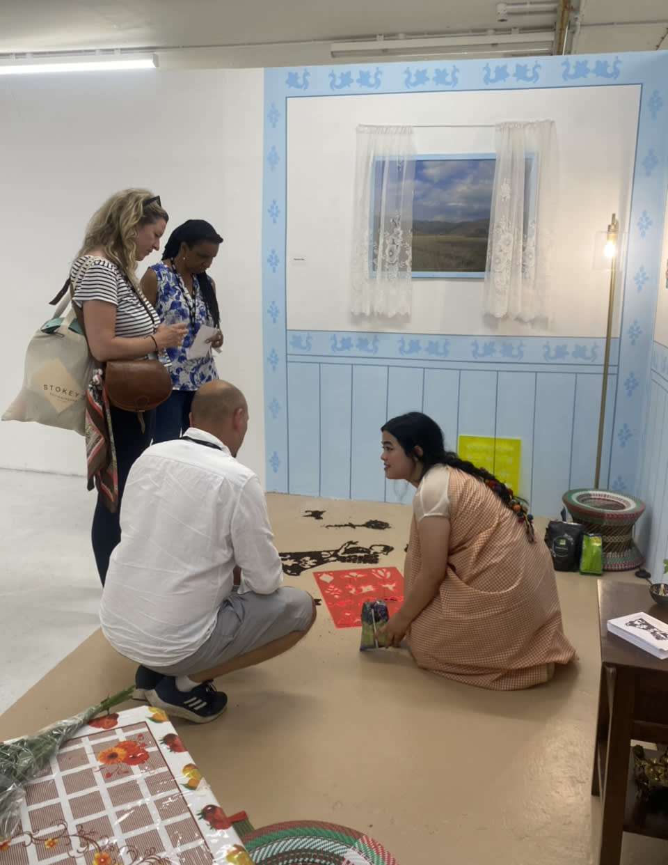 Synchar is kneeling on the floor within her installation of a domestic space, she has her dark hair tied back in a plat and is wearing a long brown dress. Another person in a white shirt crouches beside Synchar and is looking at the tea stencil and tea powder on the floor. Another two people are standing also looking at the tea powder.  