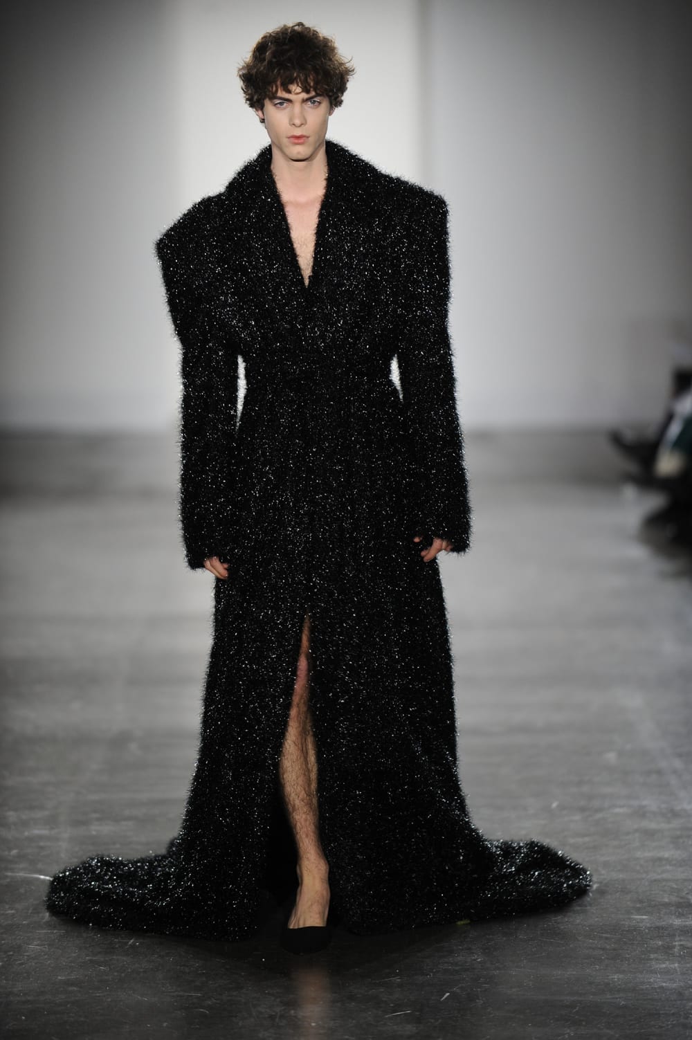 Man on runway wearing black sparkly dress with broad padded shoulder lapel collar and slit in the middle 