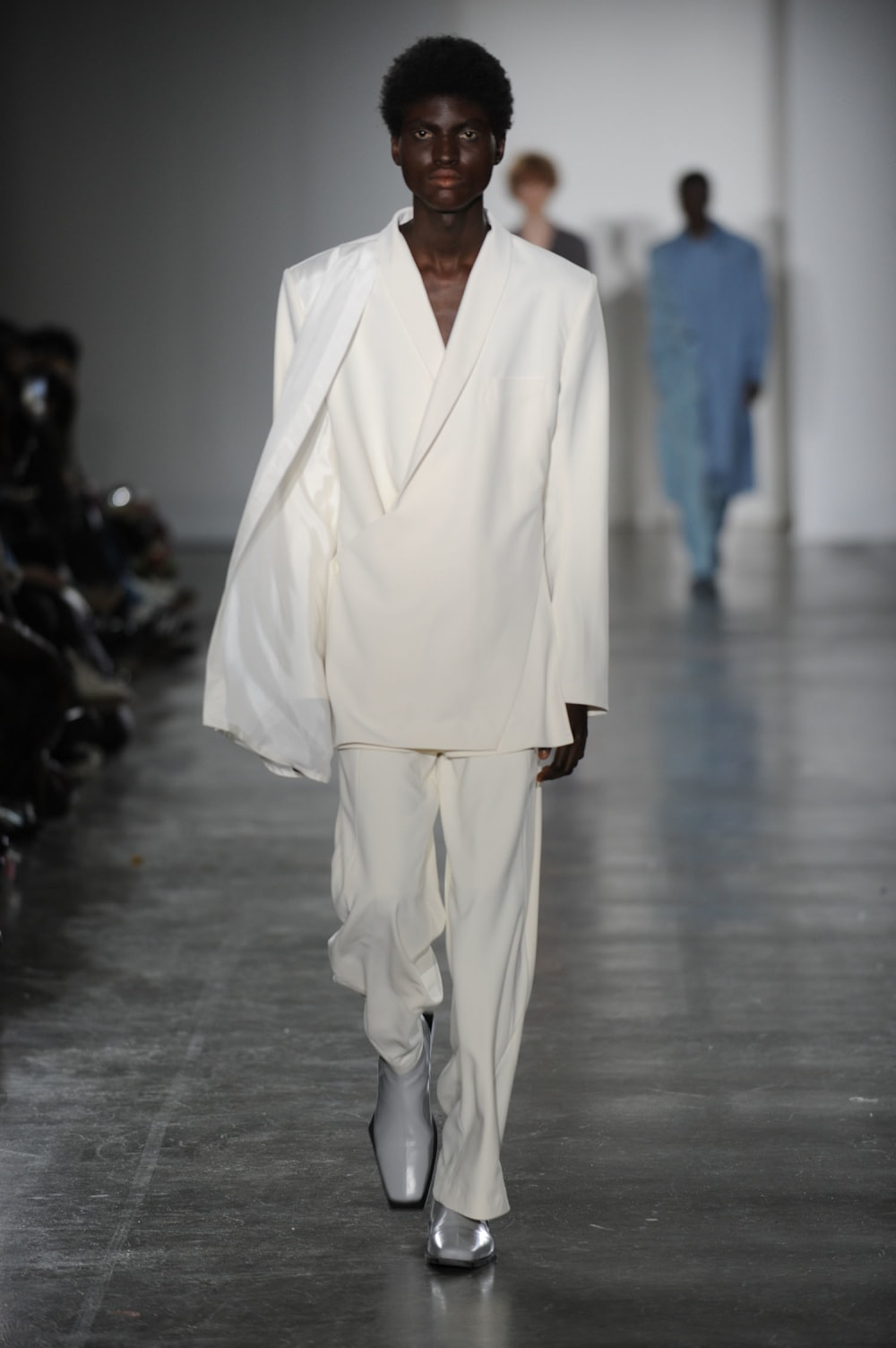 Man on runway wearing white top with padded shoulders, lapel collar and open silk material attached to right shoulder paired with white straight cut pants and white boots