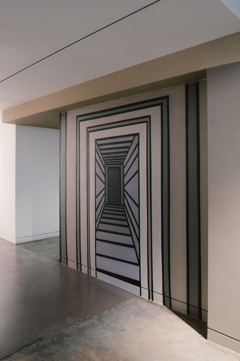 Image shows a side view of a wall with a long corridor painted in different shades of grey. The artist has used perspective to make it look as though you could walk into the corridor. There is a small black door at the end.