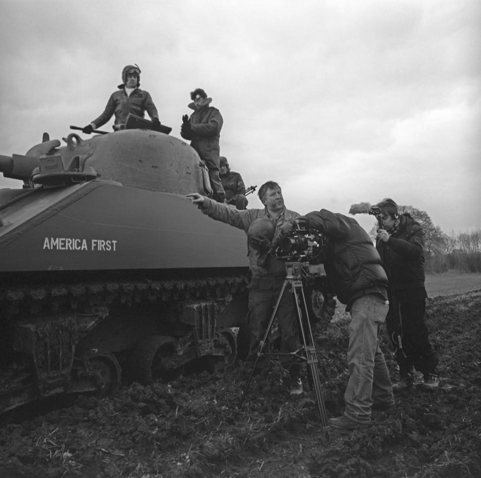Black and white photo from the film set. A World War 2 tank is in the centre, with a man in costume on top. There's 4 other men standing around who are part of the film crew