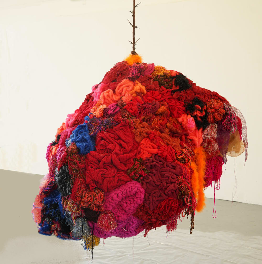 An array of folded and intertwined fabric in various colours of red, hanging from the ceiling. The cord hanging the installation is wood-like and has thorns emerging.