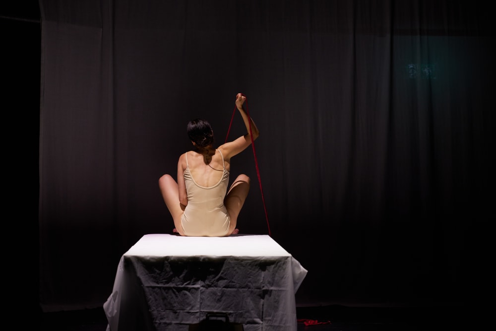 A woman in a tan bodysuit sits on a table and aggressively pulls a red string