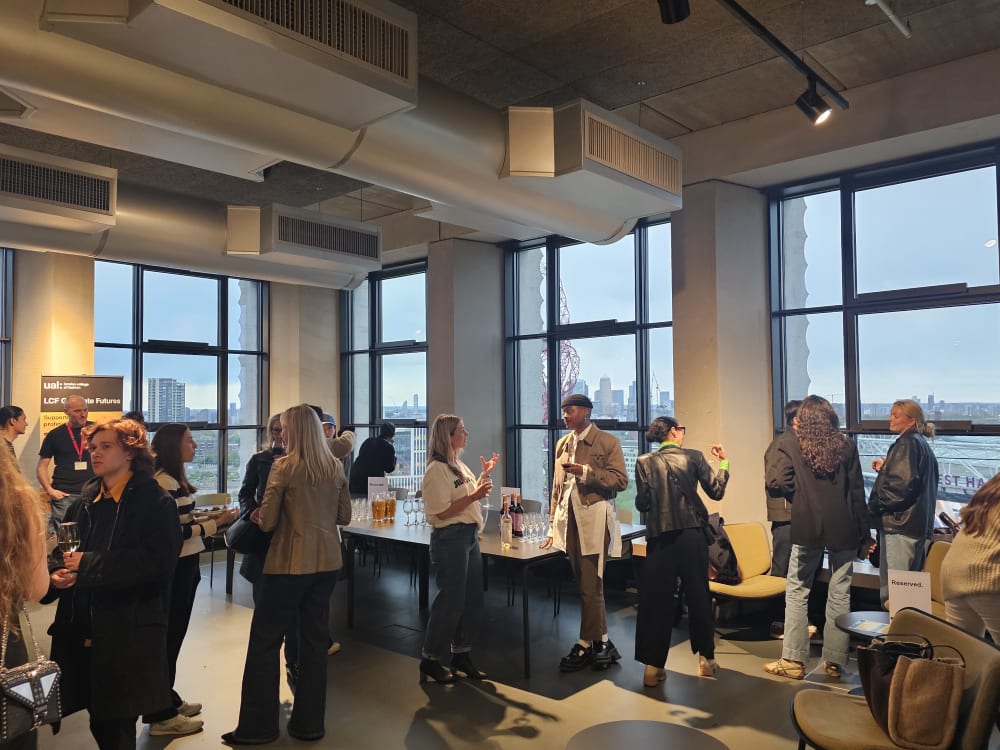 A collection of people at a networking event. Tables with food and drinks are around them, in front of tall windows. 