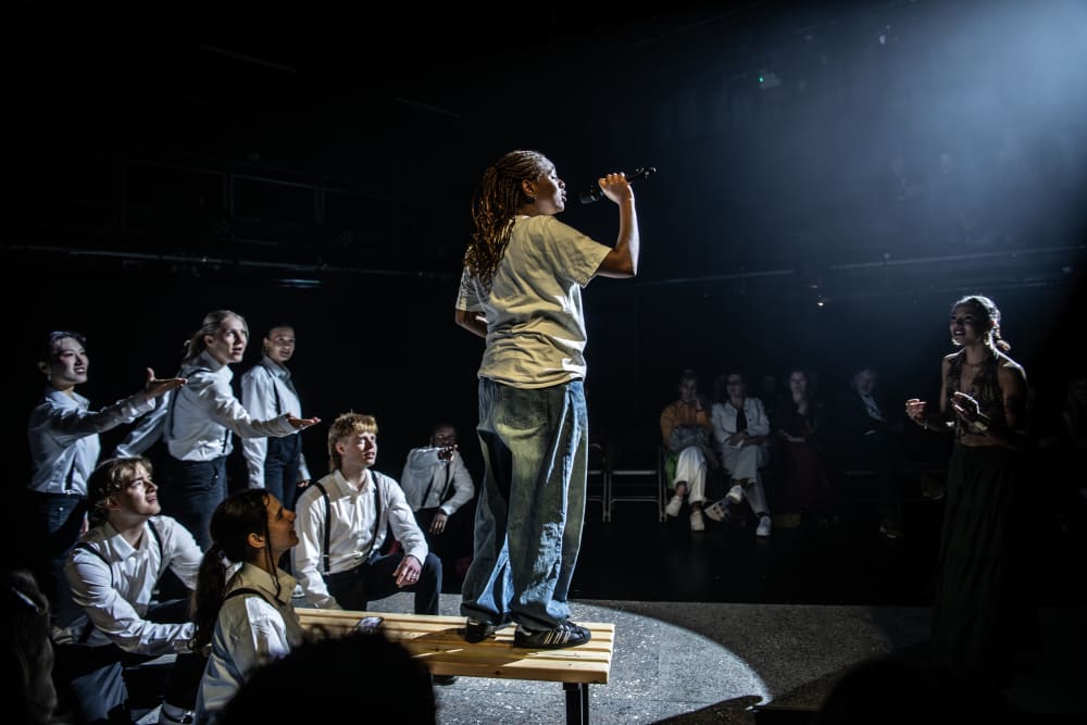 Image shows a person wearing jeans and white top standing on a wooden platform with a microphone in their hand. Around them are other people with their hands out as if to encourage the performance. 