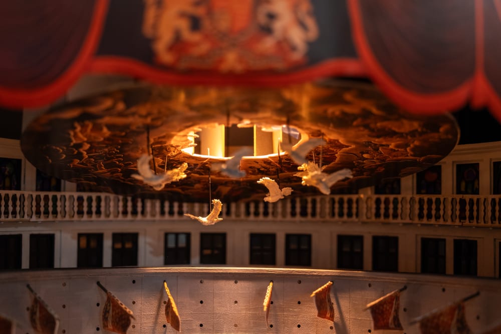 Image shows 6 small model doves circling round the roof of a building with a hole in the middle, where yellow light pours in. The image evokes a feeling of heavenlyness. 