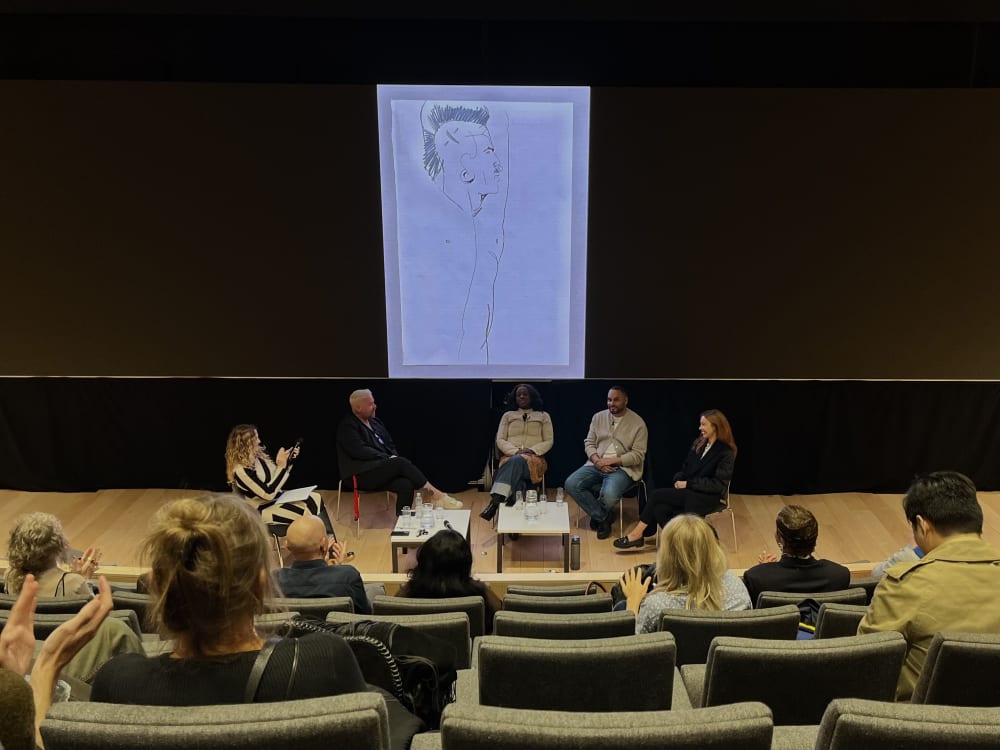 Lecture theatre with 5 people sitting at the front, and a sketch on a big screen.