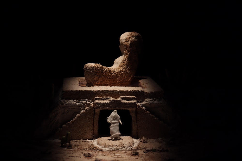 A stage design in miniature of a figure standing in front of a building made of sand. On top of the building is large Sphinx-like object. 