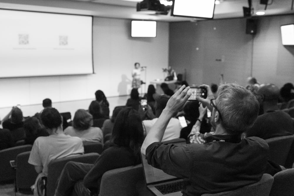 Audience sitting in lecture hall, listening to speaker at the front of hall