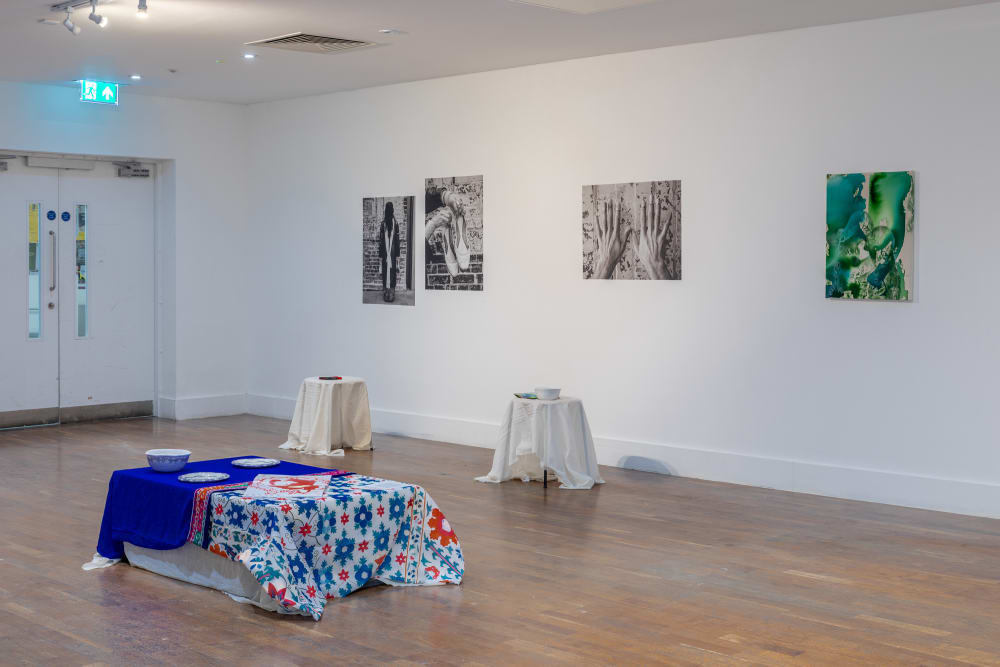 4 artworks hang on a white wall in row. Underneath are small tables with white cloth over them In the foreground is a low table covered in bright blue, white and red patterned cloth with 3 china bowls on. 
