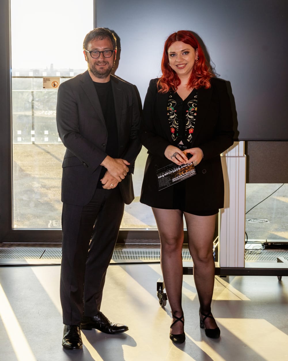 2 people standing in front of a tv screen a male on the left and a female on the right.