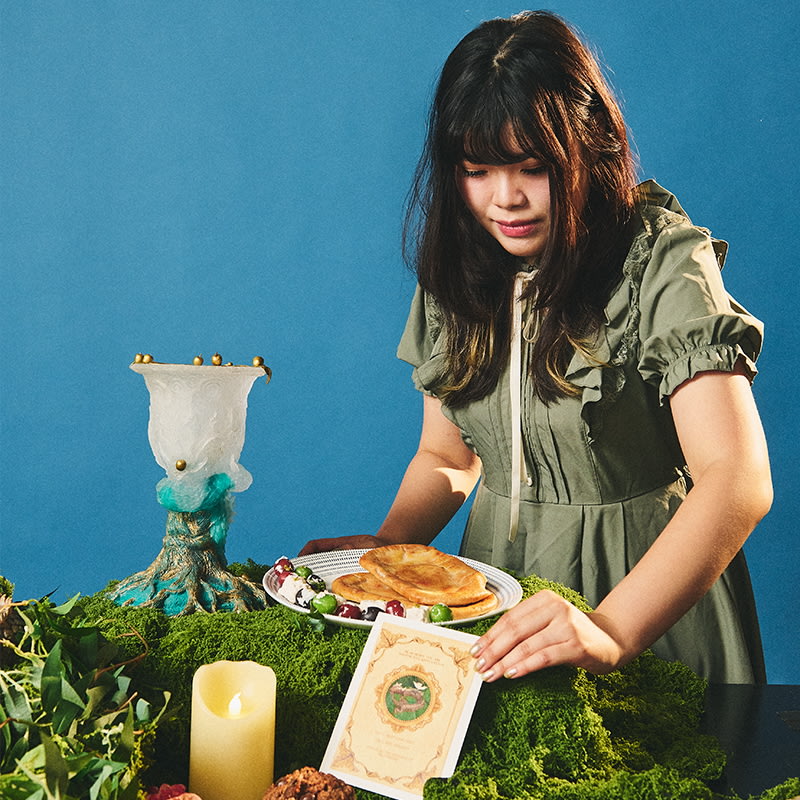 A woman in a green dress arranges a plate of bread and vegetables on a moss-covered table. A decorative candle and menu are nearby.