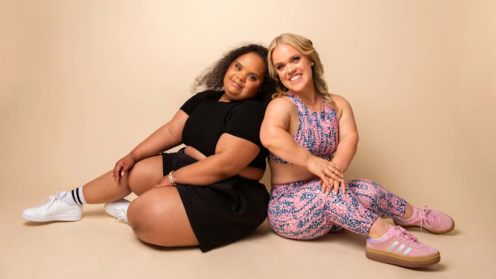 Two women sit on a light beige background. One wears a black outfit with white sneakers; the other wears a pink patterned workout set, smiling warmly.