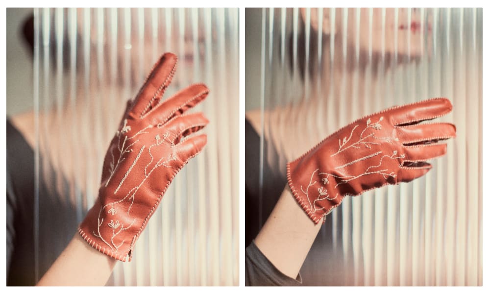 A pair of orange gloves with delicate white floral embroidery is showcased. The gloves are worn by a person partially obscured by a ribbed glass panel, creating a soft, artistic effect.
