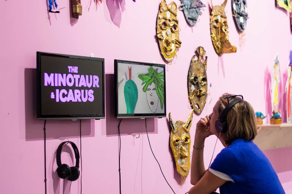 An exhibition visitor is engaging with digital artworks on 2 screens mounted on a pink wall at the Biennale. Next to the screens are large gold and silver masks and a shelf with smaller artworks.