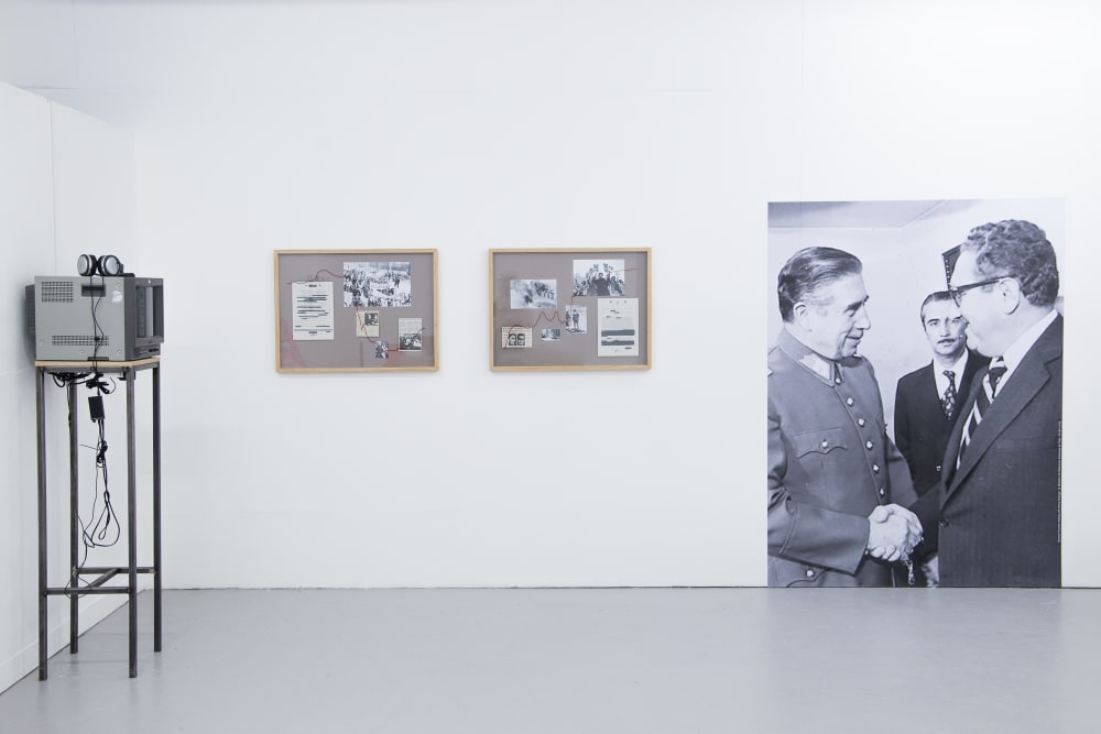 Installation view of work by BA Fine Art Photography student Ben Yau. To the left of the image is a screen on a tall stand with headphones plugged in on which a video piece will be show. To the right of this are two small framed collections of photographic images. To the right of the image is a large black and white photograph of two men shaking hands while another looks on, installed so that it touches the floor.