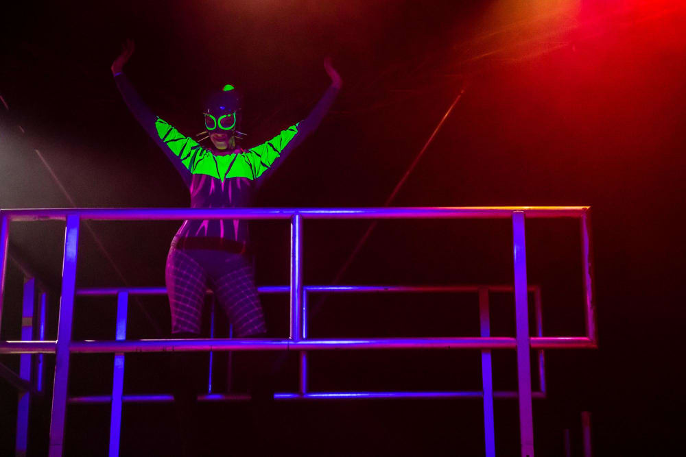 A woman in tracksuit and bobble hat stands on a scaffolding platform. Her clothes glow under UV light.