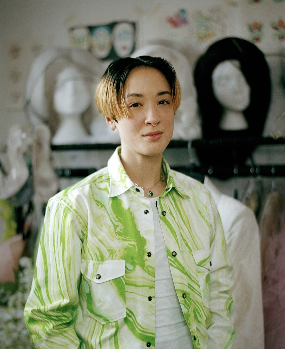 Sin Wai Kin wearing a green shirt, in the background there are wigs on mannequin heads.
