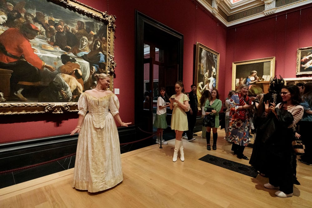 A woman in a long gold dress poses for photographs in front of a crowd of people and a large painting hanging in a grand gallery space.