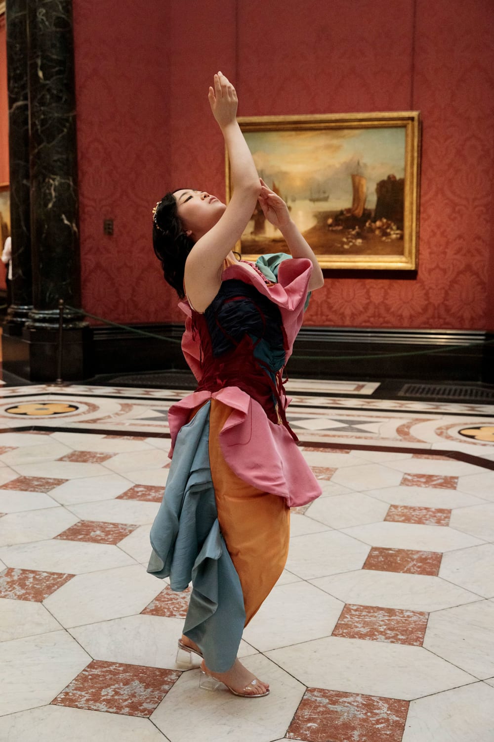 A woman in a gallery space with a marble floor poses with her arms above her head.