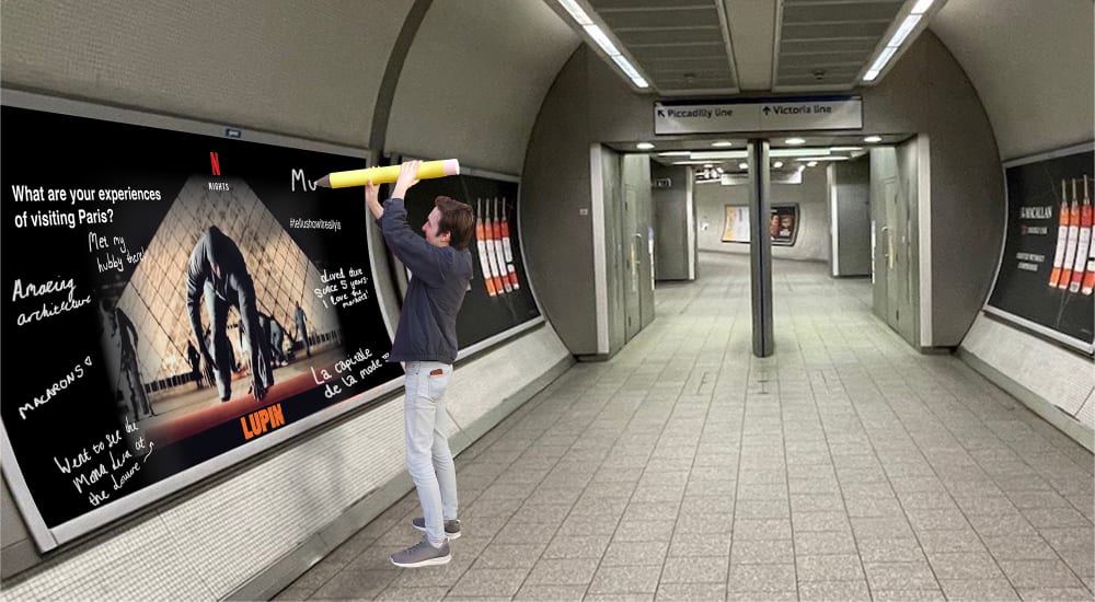 An Image of a person with a black coat on, jeans and trainers is holding a giant pencil above their head and writing on a poster positioned on the wall in an underground station. Signs for the Piccadilly Line and Victoria Line can be seen in the background.  