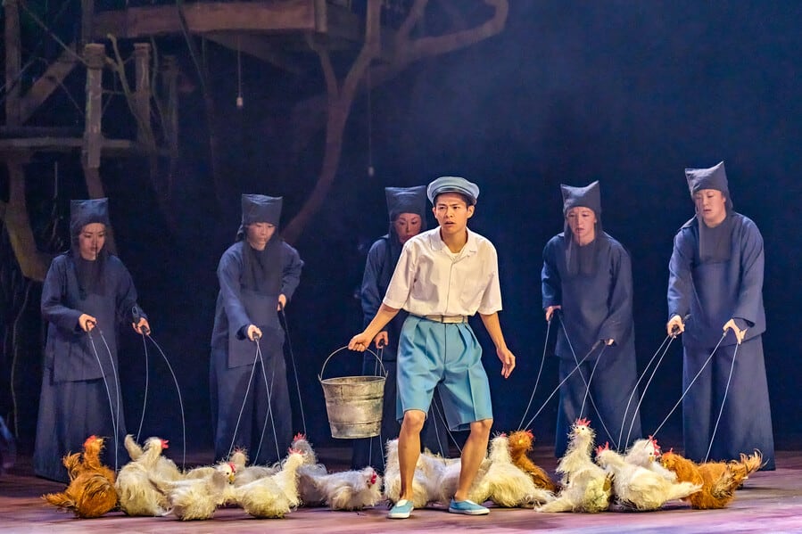 Image shows a boy with blue shorts holding a bucket that he is using to feed chickens all around his feet. The chickens are being controlled by puppeteers who are dressed in dark navy and stand in the background.