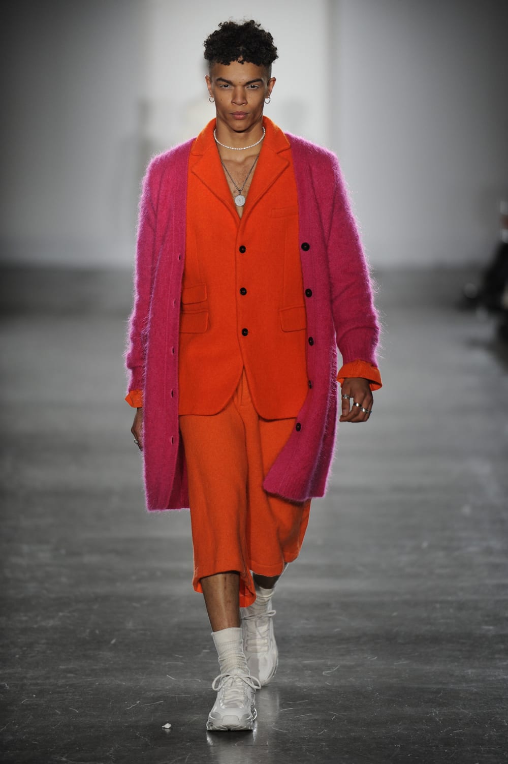 Man on runway wearing orange suit jacket with black buttons, long matching shorts paired with fuzzy pink midway cardigan with black button detail, white trainers, white long socks, silver chocker and long necklace with circle pendant and silver band rings 