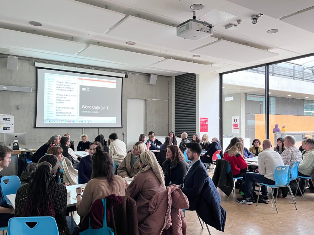 People sitting in groups in a large, well lit room with a presentation projected on a screen on the right