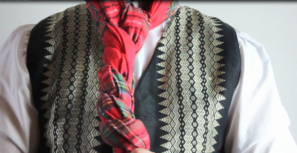 Image shows a person with a white shirt and patterned waist coat and a red tartan scarf wrapped tightly around their neck.