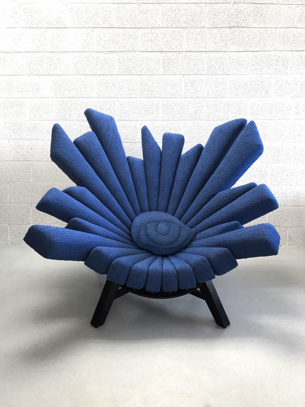 A chair with a blue hue is positioned against a white brick wall. You are drawn to the centre of the chair where there is a large eye all seeing eye. 