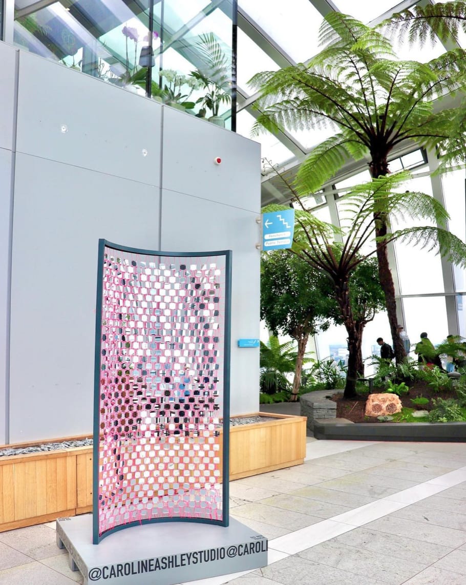 A large sculpture made of pink string and mirrored tiles sits in the foyer of the Sky Garden, a space filled with bright green plants.