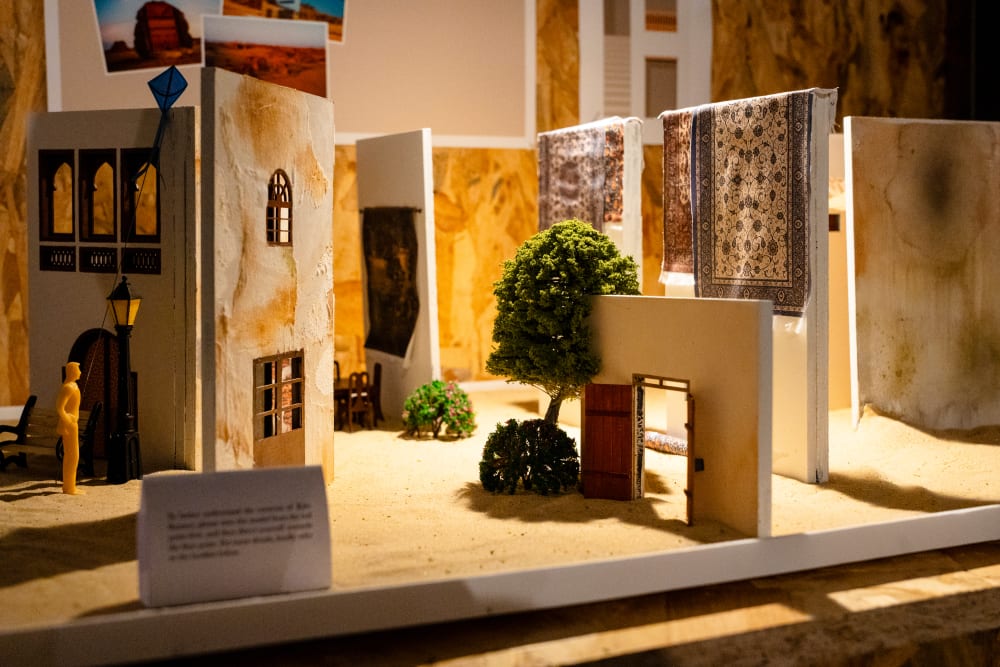 A miniature model installation of a house in a desert, with patterned carpets hanging on the walls