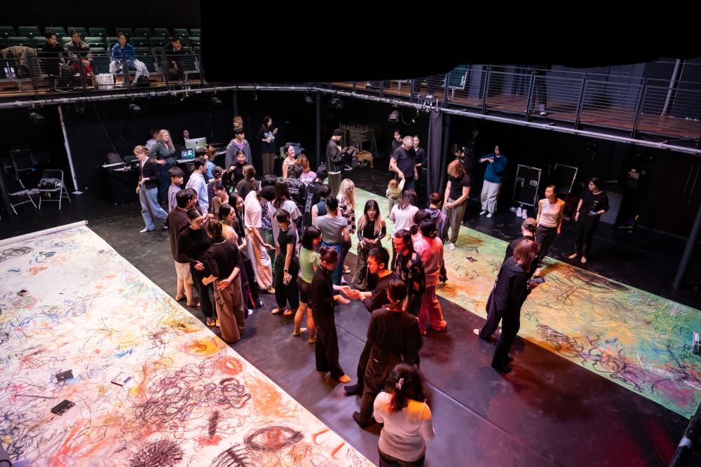 Image shows an aerial shot of a large group of people standing in a studio theatre with 2 long sheets of paper, both of which have writing and drawing on them. 