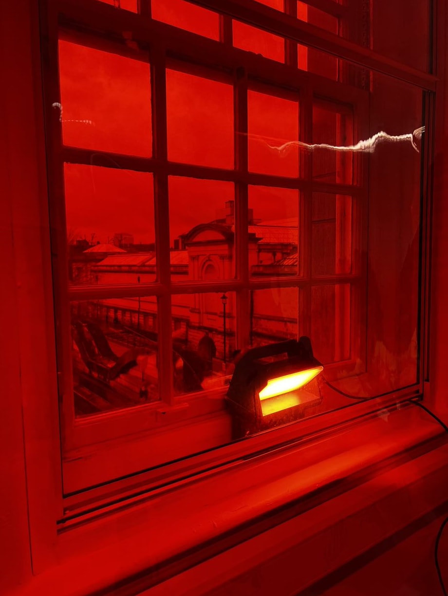 A paned window has been lit up in a deep colour of red. Through the window you can see a brick building and lamp posts.  