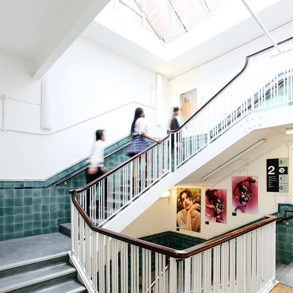 3 students walk up the staircase in the Lime Grove building for pre-degree students. There is student work on the walls.
