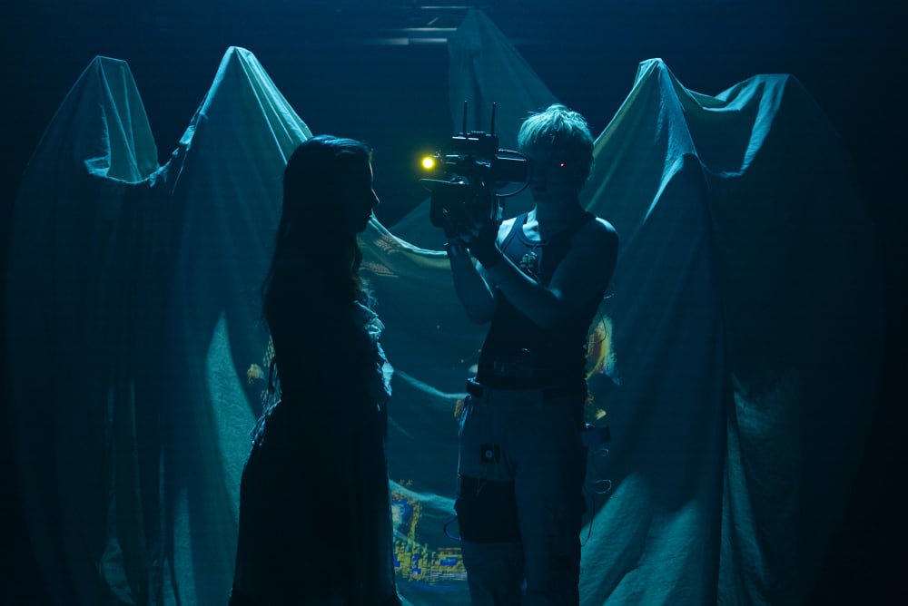 Image shows a person holding a camera filming another person. The stage is dimly lit and awash with a dark blue light.