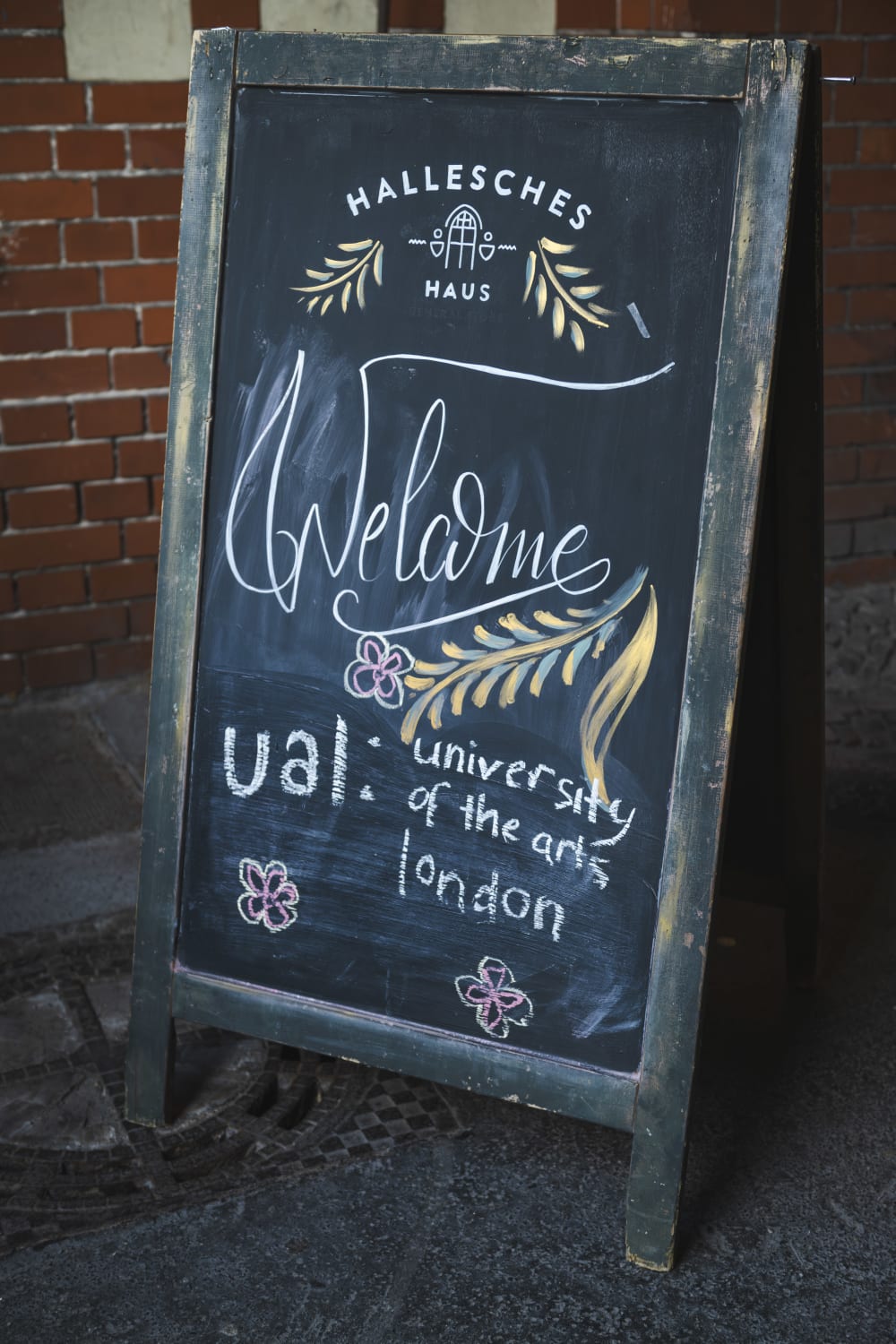 Chalkboard board sign with 'Hallsches Haus, Welcome, UAL: University of the Arts London' written on and flowers drawn around too