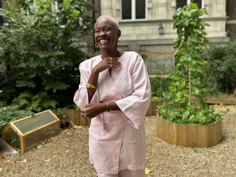 Image shows a Black woman dressed in pink laughing to the camera while standing in a garden with a grand building in the background