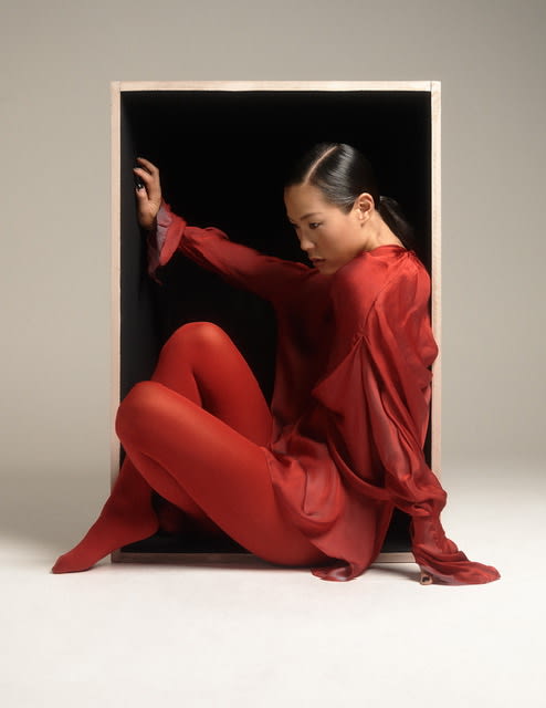 A model wearing a silk red shirt dress and red tights, sitting within a whitewashed wooden box, lying open on its side, against a white backdrop.