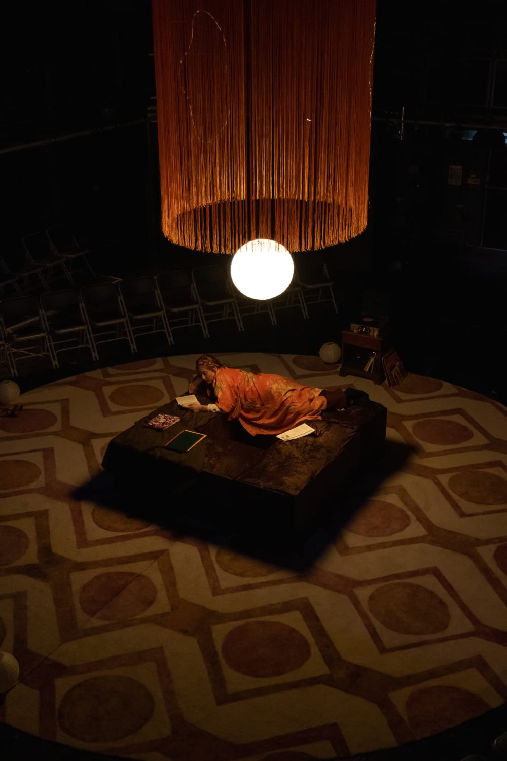 A woman is lying on a square plinth in the middle of the stage. Above her is an orb of light with orange fringing hanging above. The lighting is low.