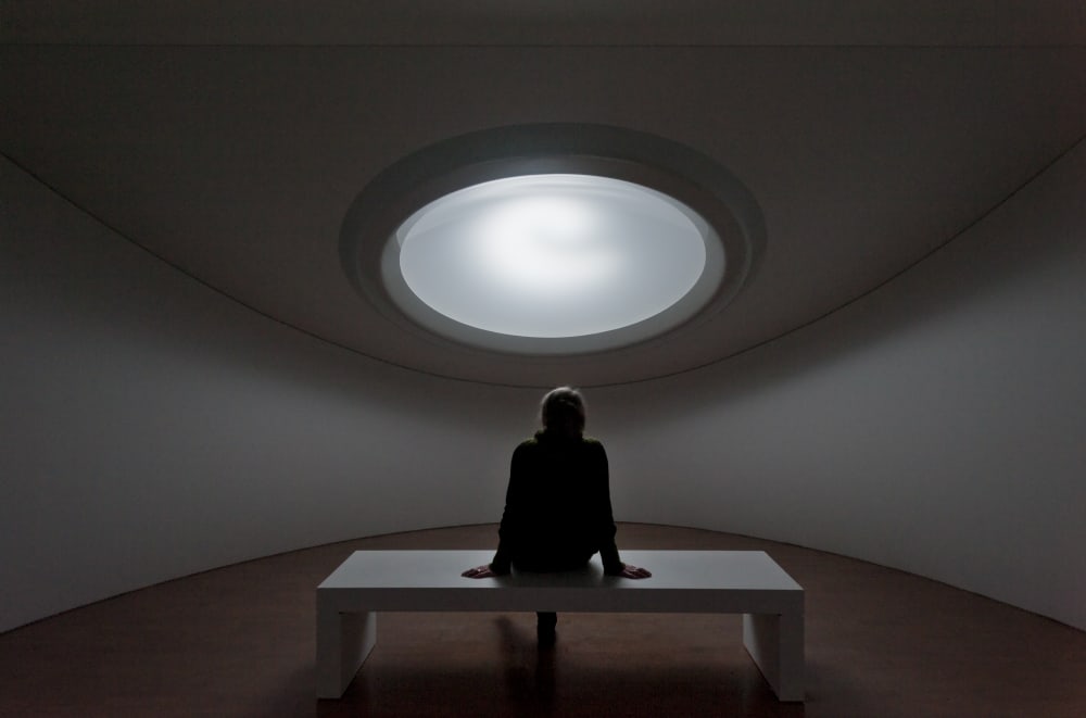  Image shows a person sitting on a bench in a room with a light red carpet, looking up towards a white hole in the ceiling. The walls in the room are also white.