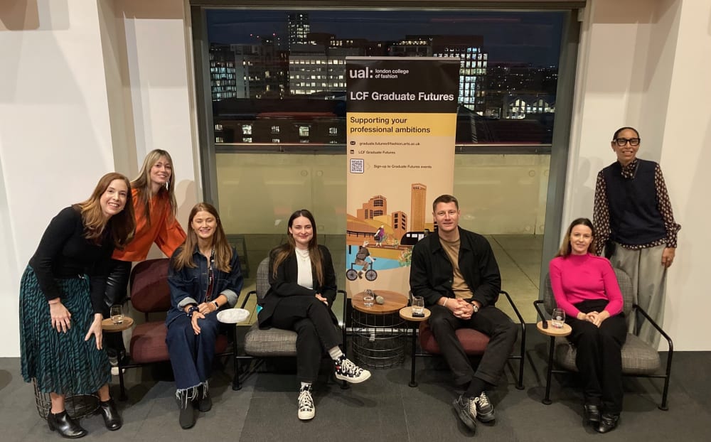7 people from the panel discussion, sitting and standing in a group in front of a window with city lights outside.