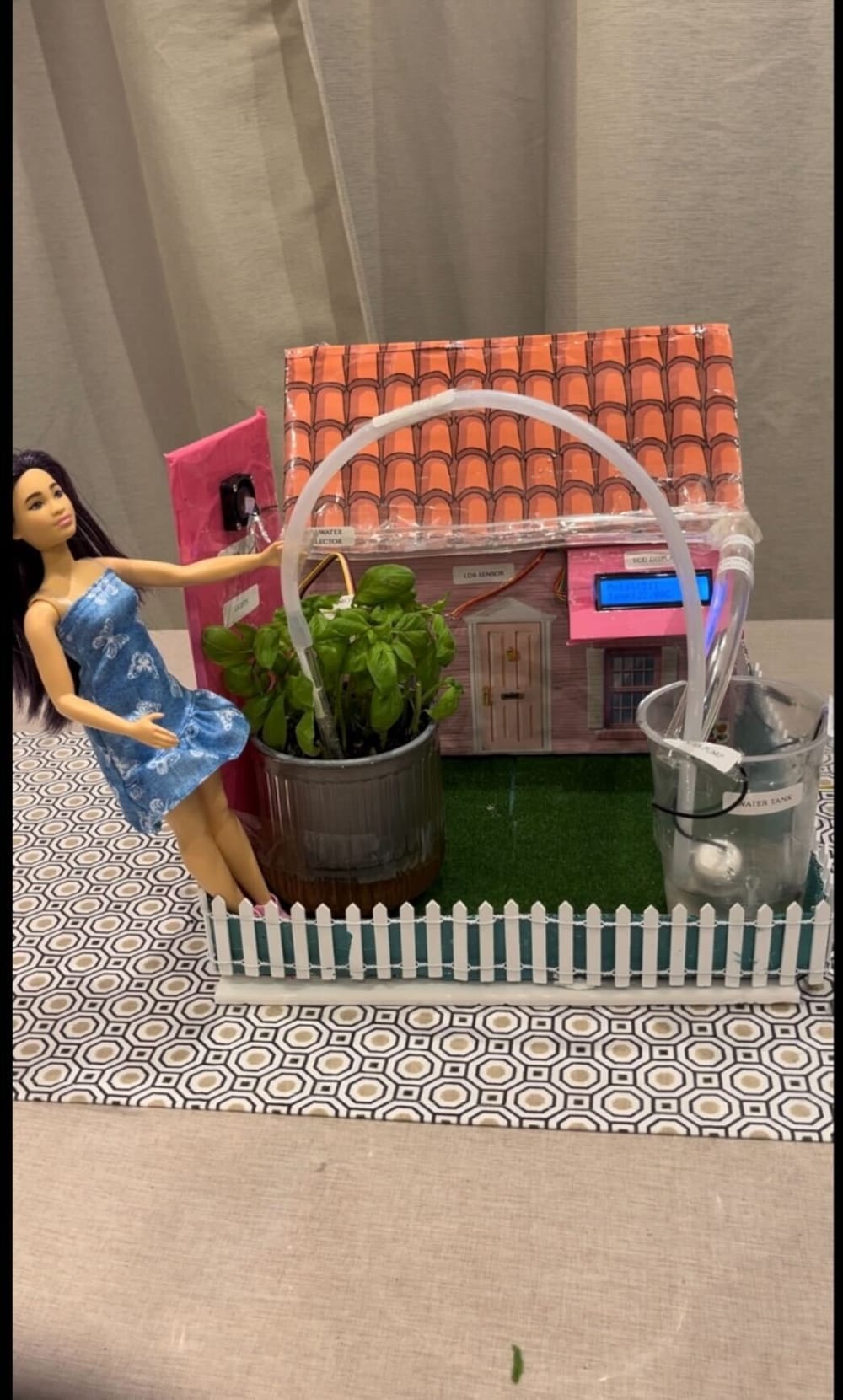 A doll in a blue dress stands beside a small model house with a red-tiled roof, surrounded by potted plants and a miniature white fence, creating a smart watering system.