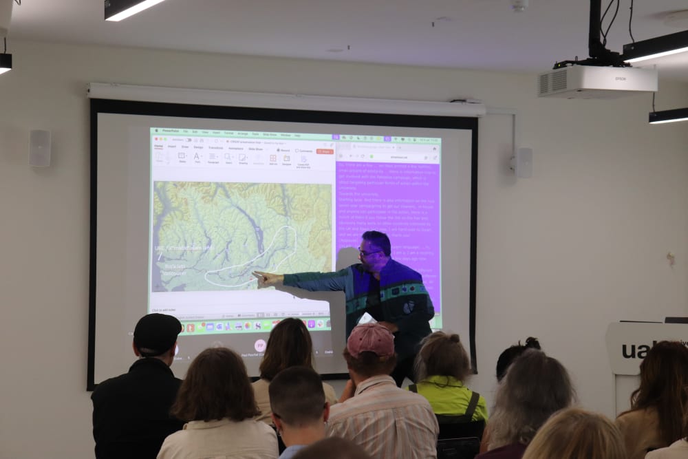 Dylan Robinson pointing at figure of a map on the projected slideshow. Backs of audience heads watching.