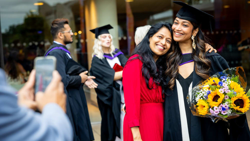 Hand holding a phone taking a picture of a Indian graduate student with her family member, with 2 graduate caucasian students in the background talking