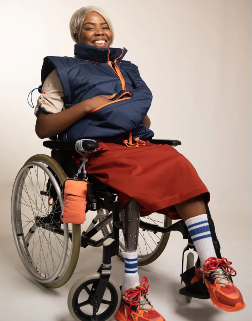 A joyful person in a wheelchair wears a blue vest, red skirt, striped socks, and bright sneakers. The image conveys a vibrant and confident mood.