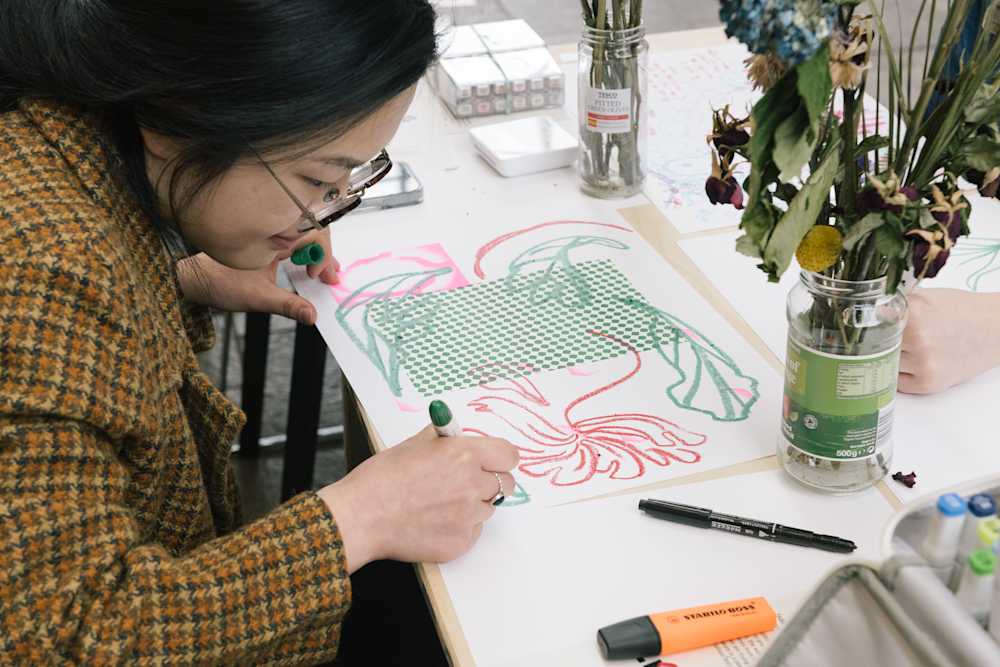 Close up on a student who is illustrating on white paper using markers and crayons on white paper. There is a glass jar with flowers in front of the paper and stationery scattered around the desk.